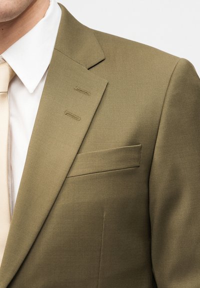 Άνδρας που φοράει σακάκι ελιάς, λευκό πουκάμισο και ανοιχτό μπεζ γραβάτα, δείχνοντας το πέτο του σακακιού, την τσέπη και τις λεπτομέρειες των ραφών.
