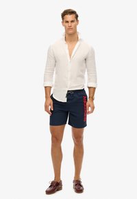Camisa de lino blanca, pantalones cortos de baño azul marino con estampado rojo "SUPERDRY", mocasines marrones, modelo de pie con el pelo corto frente a un fondo plano.
