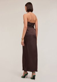 Rochie maxi din satin maro, cu decolteu halter și detalii gather la spate. Model purtând sandale verzi cu bretele. Design simplu și elegant.