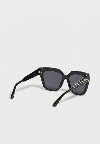 Lunettes de soleil noires à monture carrée avec verres foncés et branches noires ornées de pois blancs et d’inscriptions dorées de la marque sur un côté.