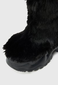 Bota de piel sintética negra con suela gruesa de goma negra. La parte superior presenta largas y suaves fibras, y una entresuela texturizada.