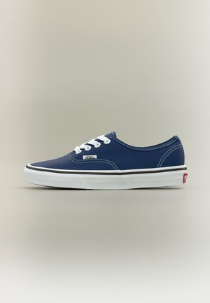 Tengerészkék Vans sportcipő fehér fűzővel, fehér talppal és Vans logóval ellátott címkével, oldalról, egyszínű háttéren bemutatva.