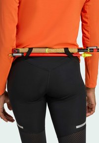 Svarta åtsittande leggings och en långärmad orange topp. Två vandringsstavar fästa i midjan med ett bälte. Reflekterande detaljer på leggings.