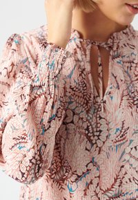 Blouse fleurie en tissu rose avec des motifs de feuilles bleues et brunes. Présente un décolleté à ouverture clé, des poignets élastiques et une surface texturée.