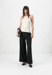 Romige, aansluitende tanktop, zwarte wijde broek en zwarte hoge hakken. Model houdt een kleine zwarte tas met een witte accent vast.