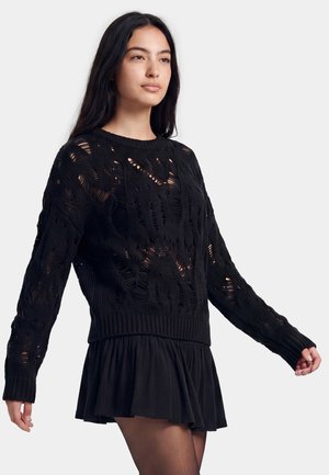 Ung kvinde med langt mørkt hår iført en sort strikket sweater med huller og en kort sort nederdel mod en ensfarvet lys baggrund.
