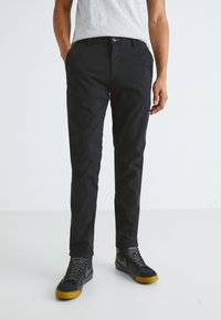 Crne hlače slim fit kroja, izrađene od glatke tkanine, s bočnim džepovima i patentnim zatvaračem, u kombinaciji s teksturiranim crnim tenisicama.