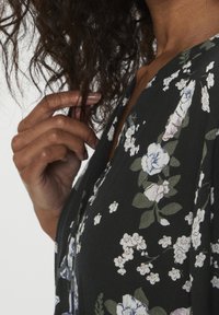 Blouse à motif floral noir, ornée de fleurs blanches et bleues, avec un décolleté en V et un tissu doux et texturé.