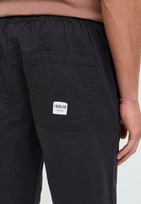 Schwarze Baumwollshorts mit elastischem Bund, ausgestattet mit einer Gesäßtasche und einem weißen rechteckigen Etikett mit dem Text "SOLID EST 1999."