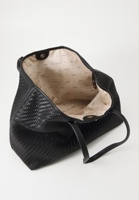 Sac fourre-tout tissé noir avec intérieur en tissu beige imprimé de logos "GUESS" et fermeture à bouton-pression magnétique, présenté ouvert avec les poignées visibles.