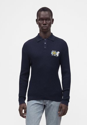 Mand iført en mørk navy tekstureret langærmet polo med patches på brystet og lyseblå jeans mod en ensfarvet hvid baggrund.