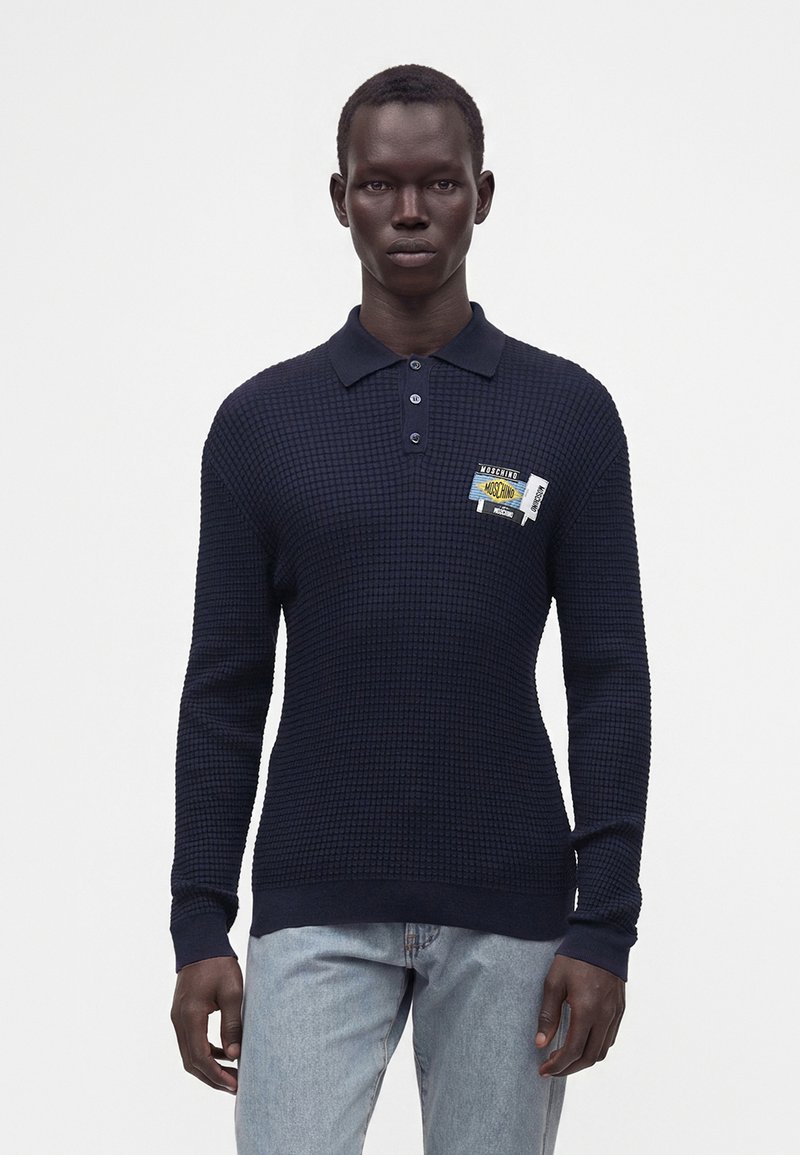 Homme portant un polo texturé bleu marine foncé à manches longues avec des écussons sur la poitrine et un jean bleu clair sur un fond blanc uni.
