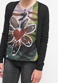 Cardigan nero sopra una maglietta a fantasia floreale multicolore con un grande fiore e un cuore rosso, in tessuto morbido, maniche corte e bottoni.