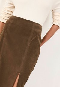 Femeie îmbrăcată cu o fustă din suede maro cu talie înaltă și o slit în față, împreună cu un top crem cu mâneci lungi, care sunt ridicate.