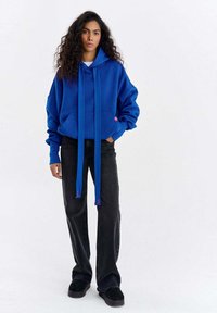 Hanorac albastru cu șnururi lungi, buzunar tip cangur, croială lejeră. Asortat cu jeansi negri, largi, și pantofi negri. Textură netedă, design casual.