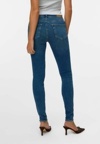 Tanke jeans v srednjem modrem denimu, z visokim pasom, dvema zadnjima žepoma in gladko teksturo. Združeni z rjavimi petami.