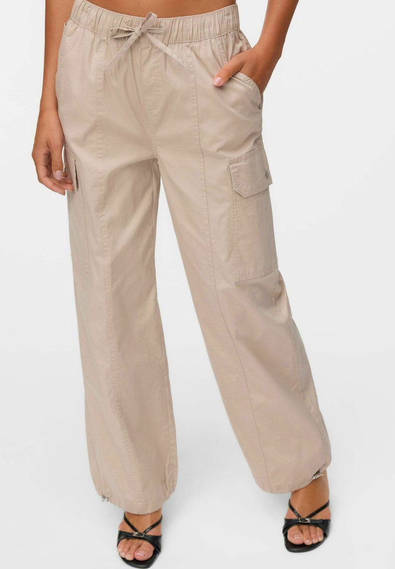 Pantalons cargo beiges avec taille élastique, cordon de serrage et poches latérales. Coupe ample avec des jambes légèrement fuselées et des accents zippés aux poignets.