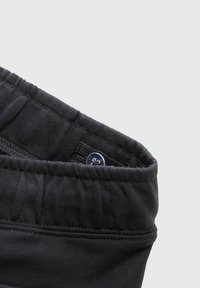 Černý elastický pás s knoflíkovým zapínáním, vyrobený z texturované látky s viditelnými švy. Celkově jednoduchý a funkční design.