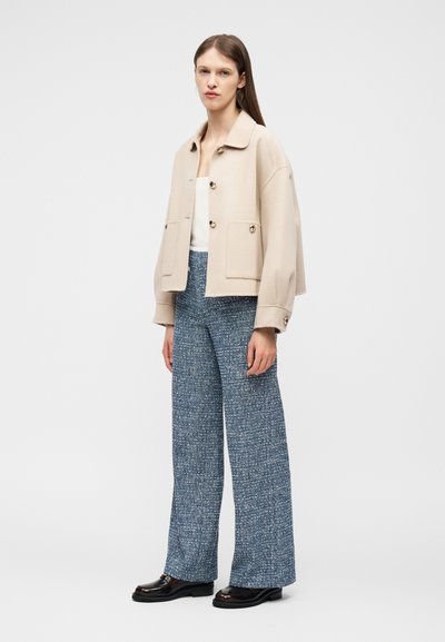 Γυναίκα με μπεζ σακάκι με κουμπιά, λευκό τοπ, μπλε παντελόνι με υφή και φαρδιά μπατζάκια, και μαύρα μοκασίνια, στέκεται μπροστά σε λευκό φόντο.