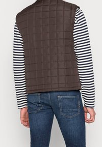 Man som bär en mörkbrun vadderad väst, en marinblå och vit randig långärmad skjorta samt blå denimjeans, vänd bort från kameran.
