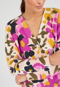 Cardigan floral avec une texture côtelée, présentant des couleurs vives comme le rose, le jaune et le vert, et un design classique à boutons avec un décolleté en V.