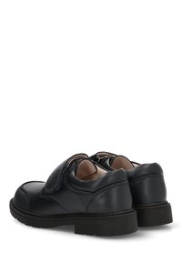 Zapatos negros de cuero para niños con tiras de velcro y suelas de goma resistentes, mostrados desde un ángulo trasero en tres cuartos sobre fondo blanco.