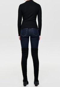 Haut noir à manches longues côtelé assorti à un jean bleu foncé et des bottes en daim noires montant au-dessus du genou avec des talons carrés. Vue de dos présentée.