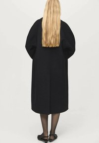 Čierny dlhý kabát s oversize rukávmi, vyrobený z hrubej tkaniny. Hladká textura, rovný strih, po kolená, s minimalistickým dizajnom.