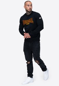 Sudadera negra con el logo "Lonsdale London", combinada con jeans negros desgastados y zapatillas blancas. El modelo posa con las manos entrelazadas.