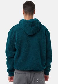 Sudadera de forro polar en color azul verdoso con una superficie texturizada, capucha y puños de canalé. Presenta un logotipo en la parte inferior izquierda. La parte delantera no es visible.