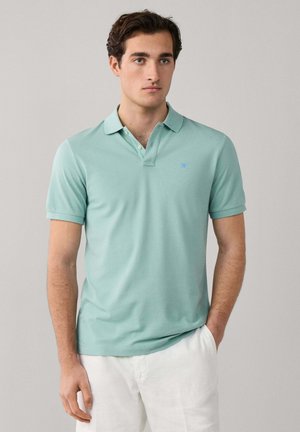 Jeune homme portant un polo manches courtes vert menthe et un pantalon blanc, debout avec une main dans la poche devant un fond uni.