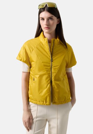 Frau mit langen dunklen Haaren trägt eine gelbe gesteppte Jacke mit kurzen Ärmeln über einem weißen Hemd und cremefarbenen Hosen, mit gelb getönten Brillen auf dem Kopf.