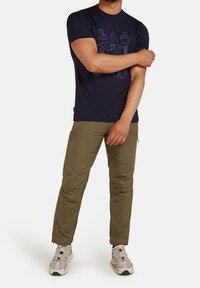 Man med marinblå grafisk t-shirt, olivgröna cargobyxor och beige sneakers, stående med ena armen korsad över den andra.