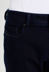 Jeans de mezclilla de color azul oscuro con un corte ajustado. Incluyen cierre de botón, bolsillo delantero con detalles en costura y una ligera textura.