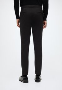 Zwarte op maat gemaakte broek met een gladde textuur, een slanke pasvorm, twee achterzakken en subtiele zijnaden. Gecombineerd met zwarte gepolijste schoenen.