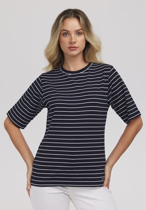 Blondynka z luźnymi falami, ubrana w granatowo-biały paskowany t-shirt z krótkim rękawem i białe spodnie, ręce na biodrach, neutralne tło.