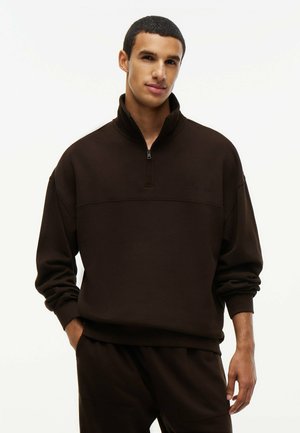 Ung mand iført en mørkebrun pullover med halvlange lynlås-krave, stående med en hånd i lommen mod en ensfarvet baggrund.