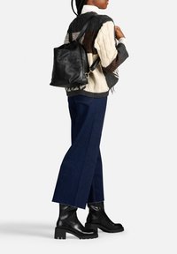 Zaino in pelle nera, maglione a trecce nei toni crema e scuri, jeans a gamba larga blu scuro, e stivali neri con tacco spesso.