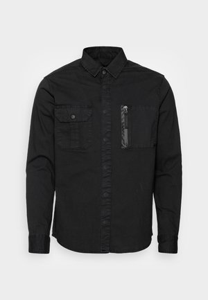 Sort langærmet skjorte med knaplukket klappocket på venstre bryst og lynlåslomme på højre bryst, spids krave og knaplukning foran.