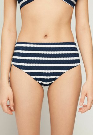 Mujer con braguita de bikini a rayas horizontales azul marino y blanca de talle alto y top a juego, mostrando el torso y las manos.