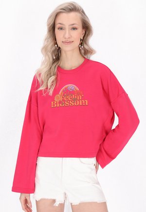 Ung kvinde med langt blondt hår iført en rød sweatshirt med teksten "Feelin' Blossom" og blomsterdesign, kombineret med hvide, frynsete shorts.