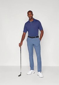 Mees seisab golfikepp käes, seljas tume sinine polo, heledad sinised püksid, must vöö ja valged spordijalatsid, ilma taustata.