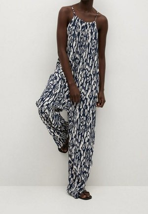 Frau trägt einen ärmellosen, locker sitzenden Jumpsuit mit einem navyblauen und weißen abstrakten Muster, stehend vor einem einfachen hellen Hintergrund.