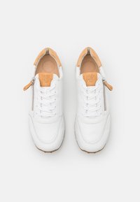 Baskets en cuir blanc avec des accents beige, présentant un bout rond, un laçage à l'avant et une fermeture éclair sur le côté pour un accès facile. Détail de semelle intérieure texturée visible.