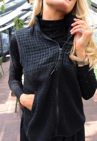 Melnas flīsa vestes ar teksturētu augšējo daļu, rāvējslēdzēja aizdare un sānu kabatas, valkātas pār pieguļošu, melnu ar traipiem garroku kreklu.