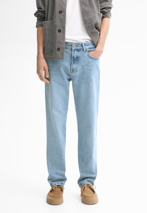 Personne portant un jean droit bleu clair, des chaussures en daim marron clair, un t-shirt blanc et une veste grise boutonnée avec les manches retroussées.