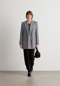 Grijze dubbele breasted wollen blazer, oversized fit, gecombineerd met een zwarte trui, donkere broek en een kleine zwarte handtas. Zachte texturen.
