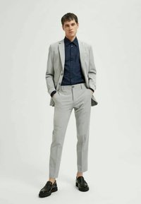 Svetlo sivi kostim z oprijetim krojem, oblečen čez temno modro srajco s subtilnim pikčastim vzorcem. Črne čevlje dopolnjujejo oblačilo.