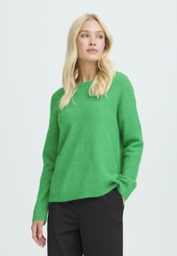 Žalias megztinis su apvaliu iškirpimu, ilgomis rankovėmis ir laisvu siluetu. Minkšta tekstūra su įlinkimo apačia ir rankovėmis. Suderintas su juodomis kelnėmis.