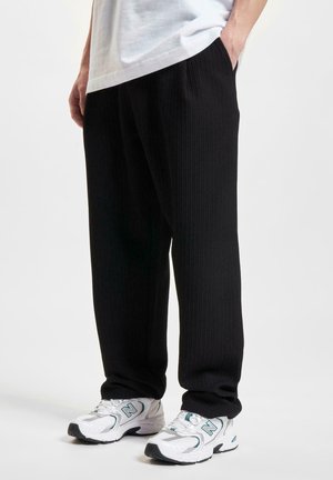 Person trägt lockere schwarze Hose und weiße New Balance Sneakers, steht mit einer Hand in der Tasche vor einem einfarbigen Hintergrund.
